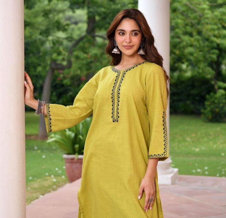 May include: A long-sleeved, chartreuse-colored tunic with black embroidered detailing along the neckline and sleeve cuffs. The tunic is made of a lightweight fabric and has a relaxed fit. The model is wearing silver earrings and bracelets.