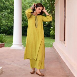 May include: A woman wearing a chartreuse-colored kurta and palazzo pants set. The long-sleeved kurta features black embroidered detailing around the neckline and cuffs. The outfit is paired with brown flats and silver earrings.