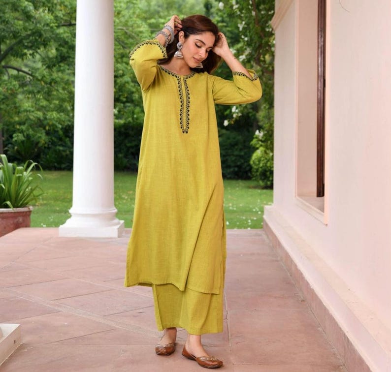 May include: A woman wearing a chartreuse-colored kurta and palazzo pants set. The long-sleeved kurta features black embroidered detailing around the neckline and cuffs. The outfit is paired with brown flats and silver earrings.