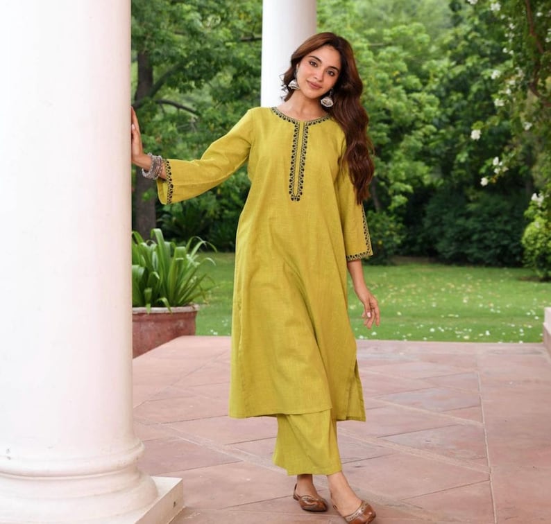 May include: A light green tunic and pants set. The tunic has long sleeves with black embroidered detailing on the neckline and cuffs. The pants are a matching light green color. The outfit is worn with brown sandals.