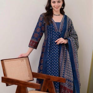 K&ouml;nnte beinhalten: Eine Frau in einem blau gemusterten Kleid und Schal. Das Kleid hat einen V-Ausschnitt und Dreiviertel&auml;rmel. Sie steht neben einem Holzstuhl mit geflochtener R&uuml;ckenlehne, der traditionelles und modernes Design vereint.