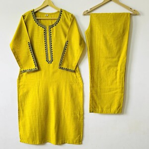 May include: A two-piece yellow outfit, consisting of a long-sleeved tunic and matching pants. The tunic features black embroidered detailing around the neckline, cuffs, and sleeves. The outfit is displayed on wooden hangers against a white background.