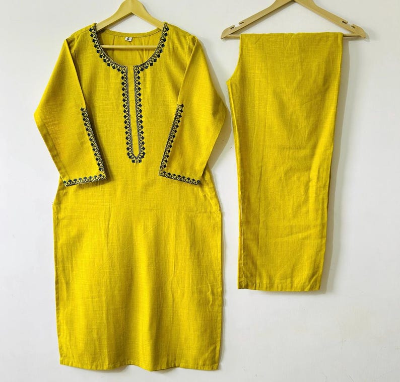 May include: A two-piece yellow outfit, consisting of a long-sleeved tunic and matching pants. The tunic features black embroidered detailing around the neckline, cuffs, and sleeves. The outfit is displayed on wooden hangers against a white background.