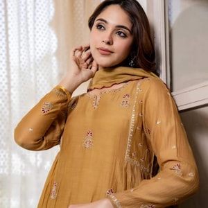 May include: Mustard-coloured long-sleeved tunic with intricate embroidery in red and white. The tunic features a round neckline and a matching scarf. The sleeves and neckline are adorned with detailed patterns. The outfit is made of a flowing fabric.