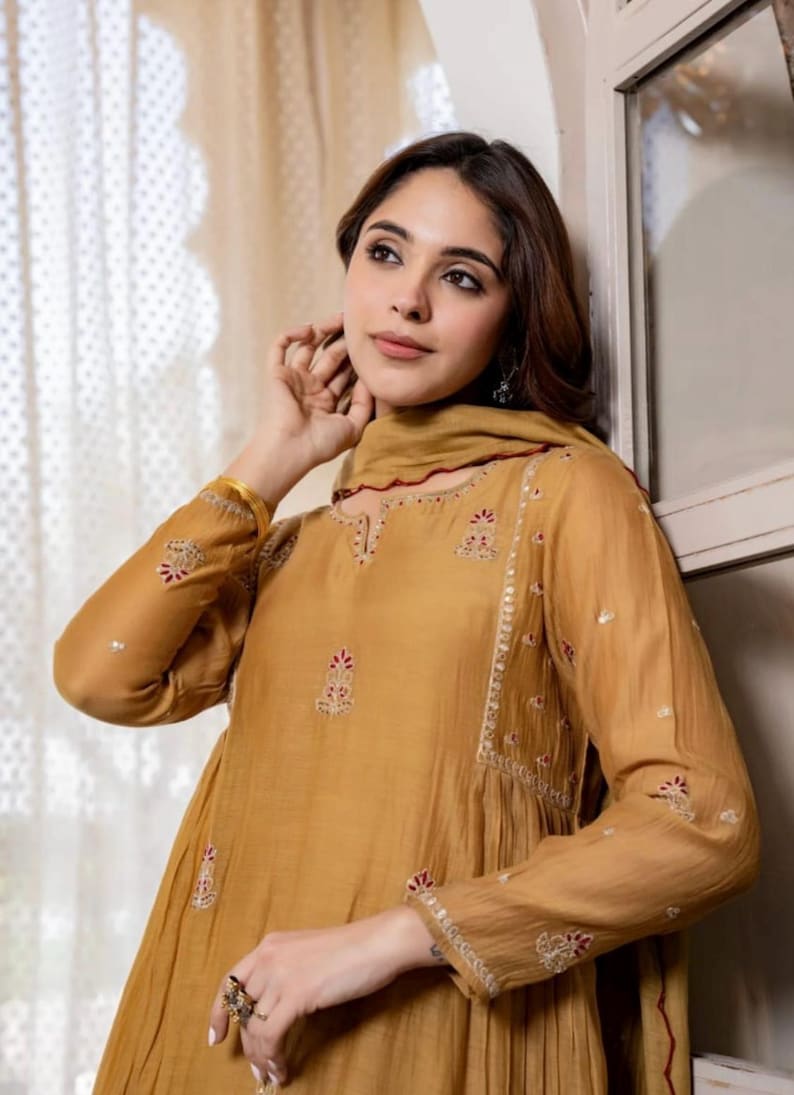 May include: Mustard-coloured long-sleeved tunic with intricate embroidery in red and white. The tunic features a round neckline and a matching scarf. The sleeves and neckline are adorned with detailed patterns. The outfit is made of a flowing fabric.