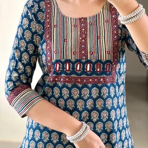 May include: A teal blue cotton kurta with a maroon and white striped neckline and a floral print. The kurta has a maroon border with silver embellishments.