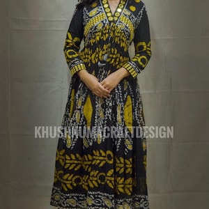 Op de afbeelding: Een lange, zwart-gele jurk met lange mouwen en een strik in de taille. De jurk heeft een wit-gouden patroon en is versierd met kleine ronde spiegels. De jurk wordt gedragen door een persoon met lang donker haar. De tekst "KHUSHNUMACRAFTDESIGN" is zichtbaar onderaan de afbeelding.