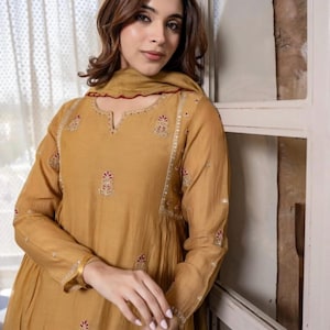 May include: Mustard-coloured long-sleeve dress with intricate embroidery and a matching scarf. The dress features a V-neckline and delicate floral patterns in red and white. The sleeves are long and the dress has a flowing silhouette.