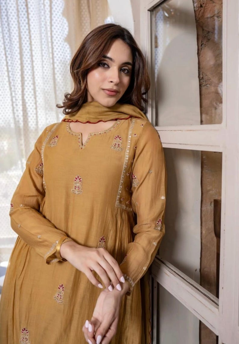 May include: Mustard-coloured long-sleeve dress with intricate embroidery and a matching scarf. The dress features a V-neckline and delicate floral patterns in red and white. The sleeves are long and the dress has a flowing silhouette.