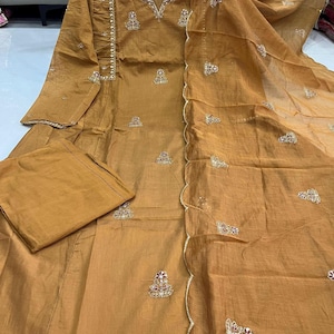 May include: Mustard-coloured salwar kameez set with embroidery. The outfit comprises a long-sleeved tunic, matching trousers, and a sheer dupatta. The tunic has detailed embroidery and embellishments on the neckline and sleeves. The dupatta has a scalloped edge.
