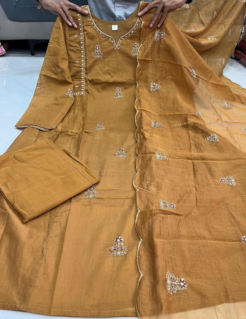 May include: Mustard-coloured salwar kameez set with embroidery. The outfit comprises a long-sleeved tunic, matching trousers, and a sheer dupatta. The tunic has detailed embroidery and embellishments on the neckline and sleeves. The dupatta has a scalloped edge.