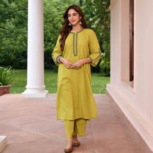 May include: A woman wearing a lime green kurta and matching pants. The kurta has black embroidered detailing around the neckline and cuffs. The outfit is paired with brown flats. The background features a garden setting with a white column.