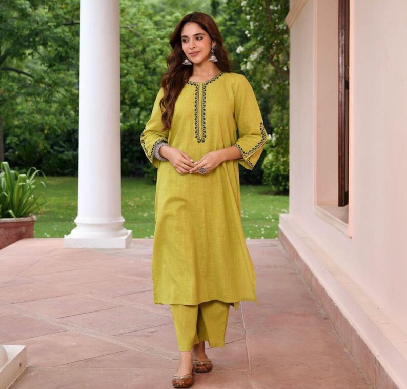 May include: A woman wearing a lime green kurta and matching pants. The kurta has black embroidered detailing around the neckline and cuffs. The outfit is paired with brown flats. The background features a garden setting with a white column.