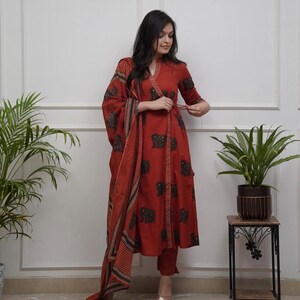 May include: A rust-colored, wrap-style kurta suit with a matching dupatta. The kurta and pants feature a black elephant print. The dupatta has a patterned border. The outfit is worn with gold-colored sandals.