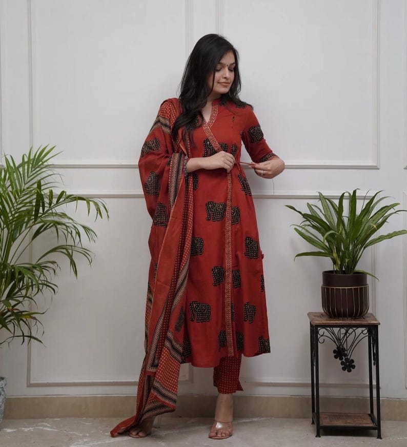 May include: A rust-colored, wrap-style kurta suit with a matching dupatta. The kurta and pants feature a black elephant print. The dupatta has a patterned border. The outfit is worn with gold-colored sandals.