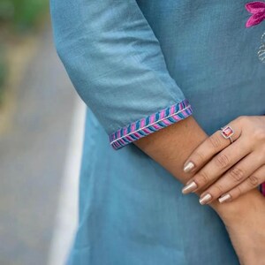 Peut inclure: Une tunique en coton bleu clair avec des bordures brod&eacute;es roses et bleues sur les manches. La tunique a un effet ombr&eacute; subtil.