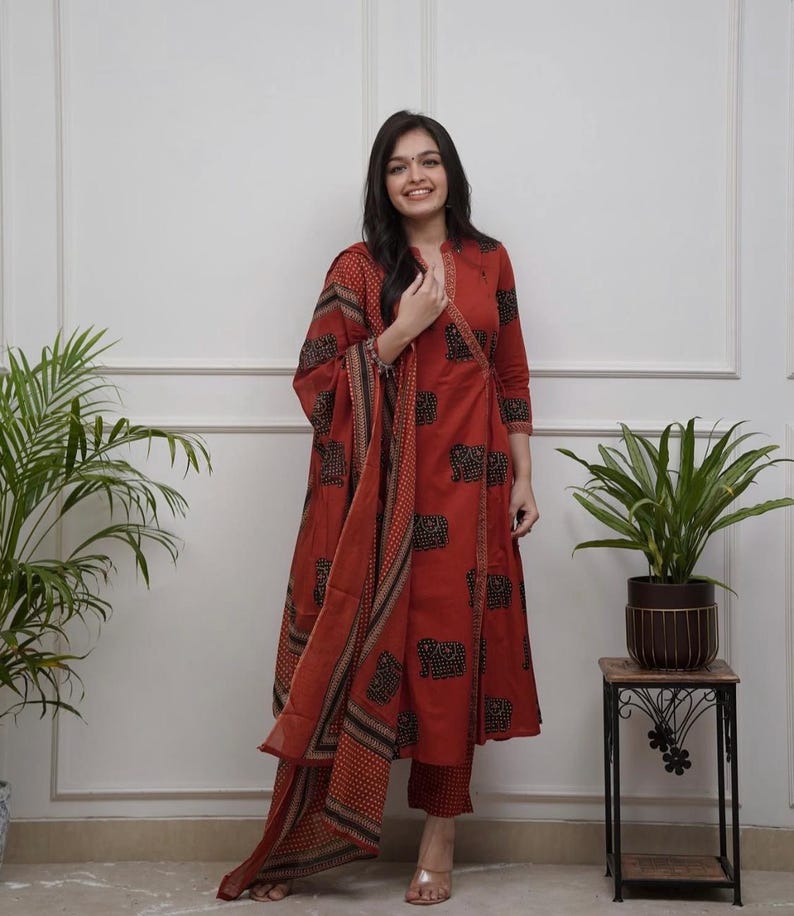 May include: A rust-colored, three-piece Indian suit featuring a long-sleeved tunic, matching pants, and a patterned scarf. The tunic and scarf have a repeating black and tan elephant design. The outfit is worn with clear sandals.