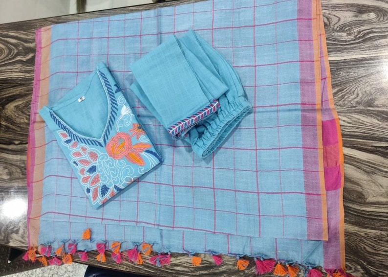 Peut inclure: Un ensemble salwar kameez en coton bleu clair avec des d&eacute;tails floraux brod&eacute;s. L'ensemble comprend un haut &agrave; manches longues, un pantalon et une dupatta assortie &agrave; motif &agrave; carreaux et des glands roses et orange.