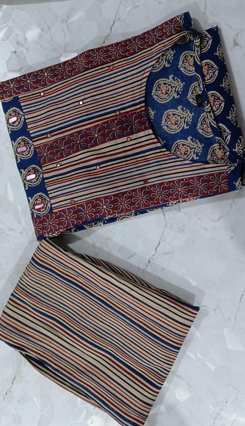 May include: A two-piece clothing set featuring a striped top with a floral border and a matching striped bottom. The top has a round neckline and is decorated with small, shiny embellishments. The fabric is a light brown with blue, red, and black stripes.