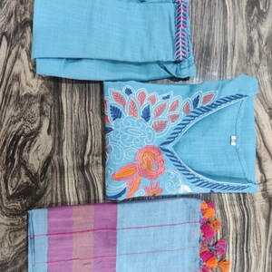 Peut inclure: Un ensemble de v&ecirc;tements trois pi&egrave;ces bleu clair avec des broderies florales. L'ensemble comprend un haut &agrave; manches longues, un pantalon et une &eacute;charpe assortie avec des glands.