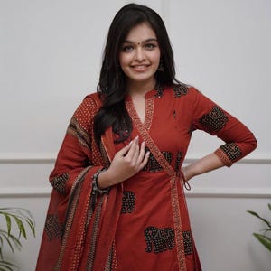 May include: A rust-colored wrap dress and matching scarf with a black elephant print. The dress has long sleeves and a decorative trim. The outfit is a traditional style.