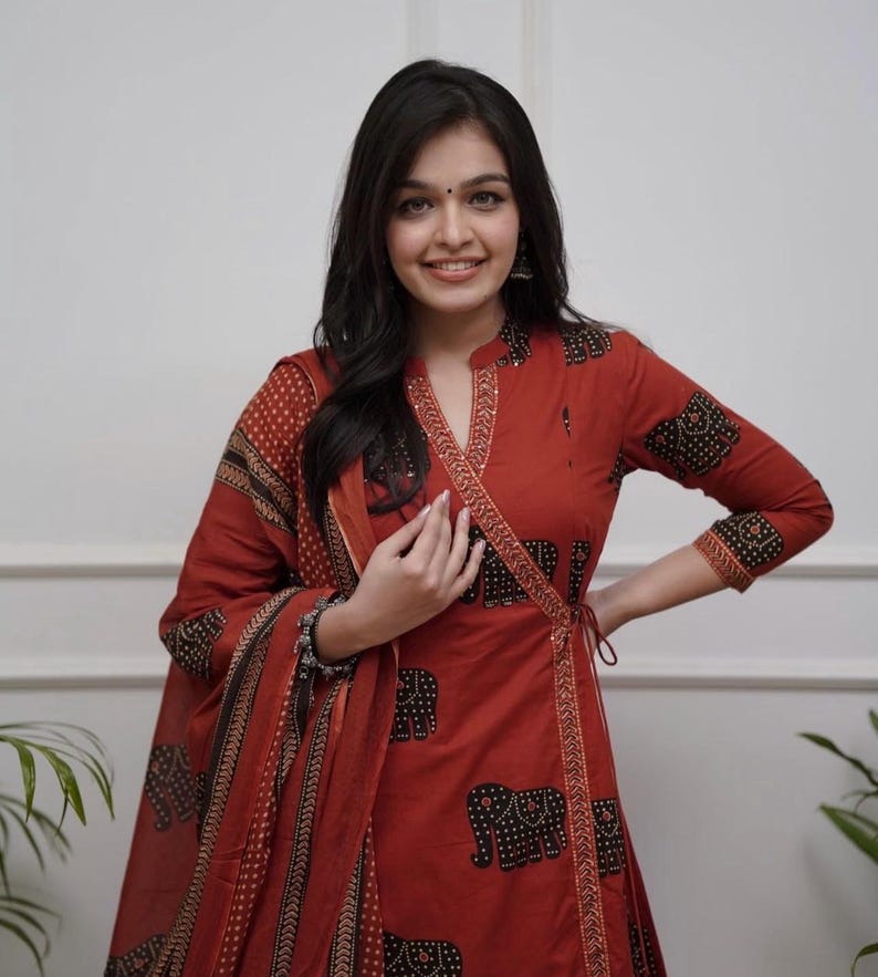 May include: A rust-colored wrap dress and matching scarf with a black elephant print. The dress has long sleeves and a decorative trim. The outfit is a traditional style.