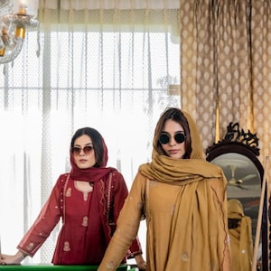 May include: Two people in matching outfits and sunglasses stand near a green pool table. One wears a maroon outfit, the other a gold-coloured outfit. Both outfits have long sleeves and decorative embroidery. A cue stick leans against a mirror.