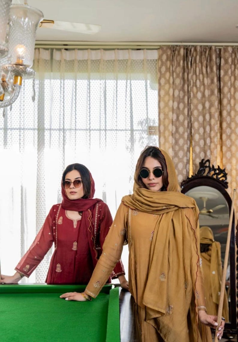 May include: Two people in matching outfits and sunglasses stand near a green pool table. One wears a maroon outfit, the other a gold-coloured outfit. Both outfits have long sleeves and decorative embroidery. A cue stick leans against a mirror.