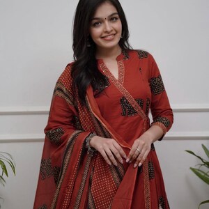 May include: A rust-colored Indian suit with a wrap-style top and matching dupatta. The suit features a black elephant print and a dotted pattern. The sleeves are three-quarter length. The outfit is worn by a person.