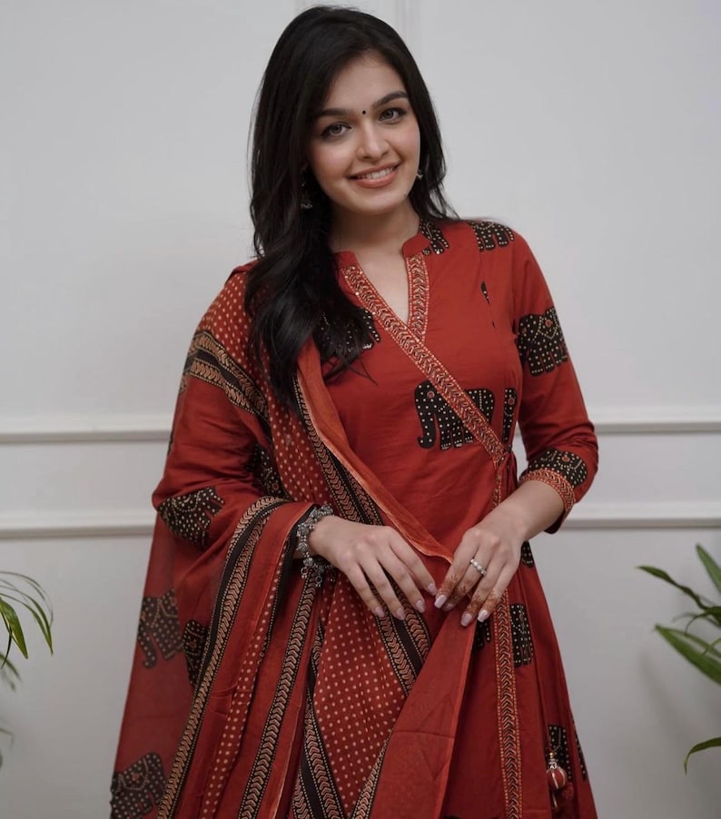 May include: A rust-colored Indian suit with a wrap-style top and matching dupatta. The suit features a black elephant print and a dotted pattern. The sleeves are three-quarter length. The outfit is worn by a person.