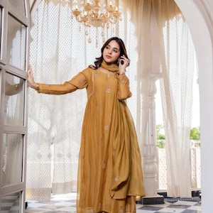 May include: Mustard-coloured, embroidered tunic dress with matching trousers and scarf. The long-sleeved dress features delicate embroidery and a flowing silhouette. The outfit is worn with clear sandals. A chandelier hangs in the background.