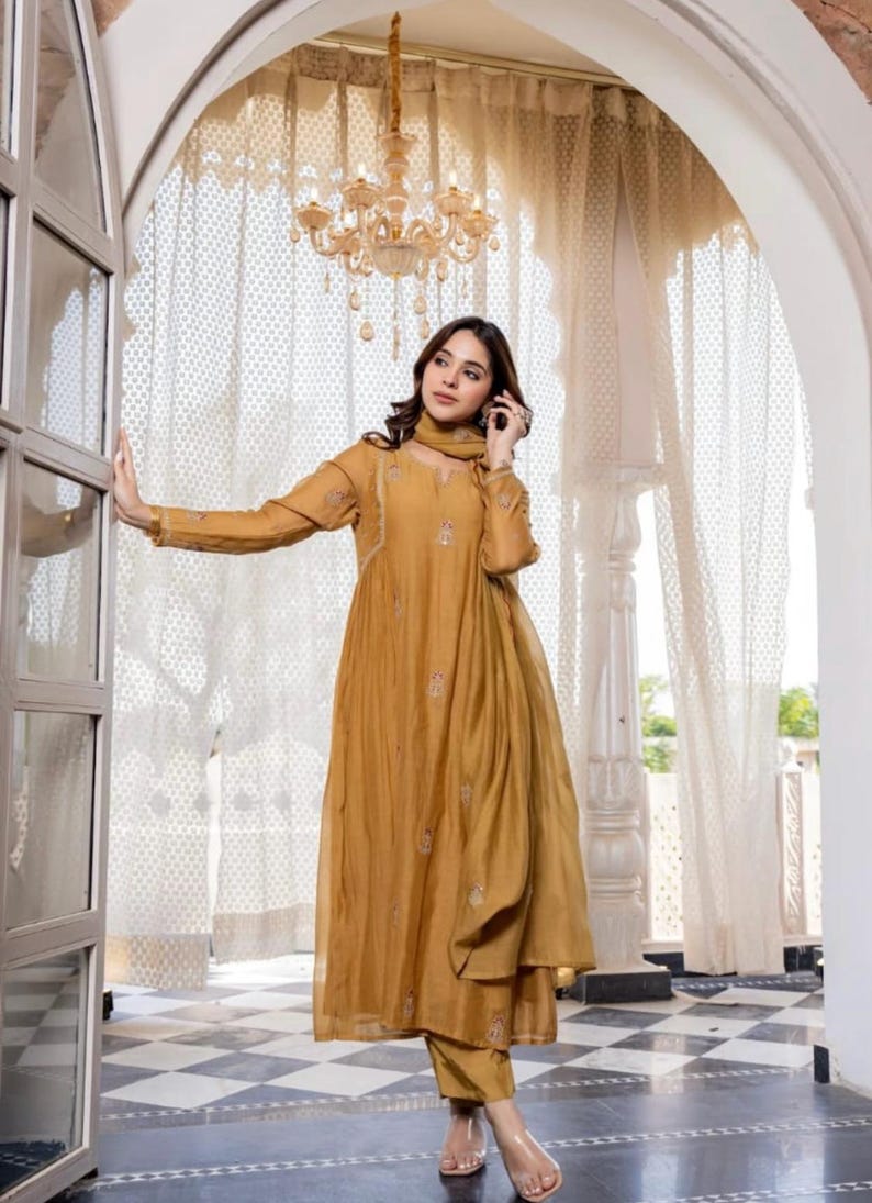 May include: Mustard-coloured, embroidered tunic dress with matching trousers and scarf. The long-sleeved dress features delicate embroidery and a flowing silhouette. The outfit is worn with clear sandals. A chandelier hangs in the background.