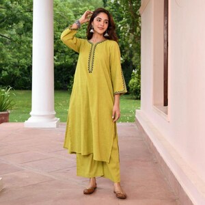 May include: A woman wearing a lime green kurta and matching palazzo pants. The kurta has black embroidered detailing around the neckline and sleeve cuffs. The outfit is paired with brown flats. The background includes a white column and greenery.