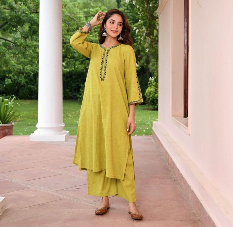 May include: A woman wearing a lime green kurta and matching palazzo pants. The kurta has black embroidered detailing around the neckline and sleeve cuffs. The outfit is paired with brown flats. The background includes a white column and greenery.