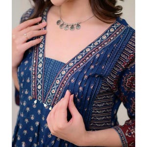 K&ouml;nnte beinhalten: Ein blau gemustertes Kleid mit V-Ausschnitt und Dreiviertel&auml;rmeln. Das Kleid hat ein detailliertes Design mit Falten und dekorativen Verzierungen. Eine silberne Halskette mit kreisf&ouml;rmigen Anh&auml;ngern wird mit dem Kleid getragen.