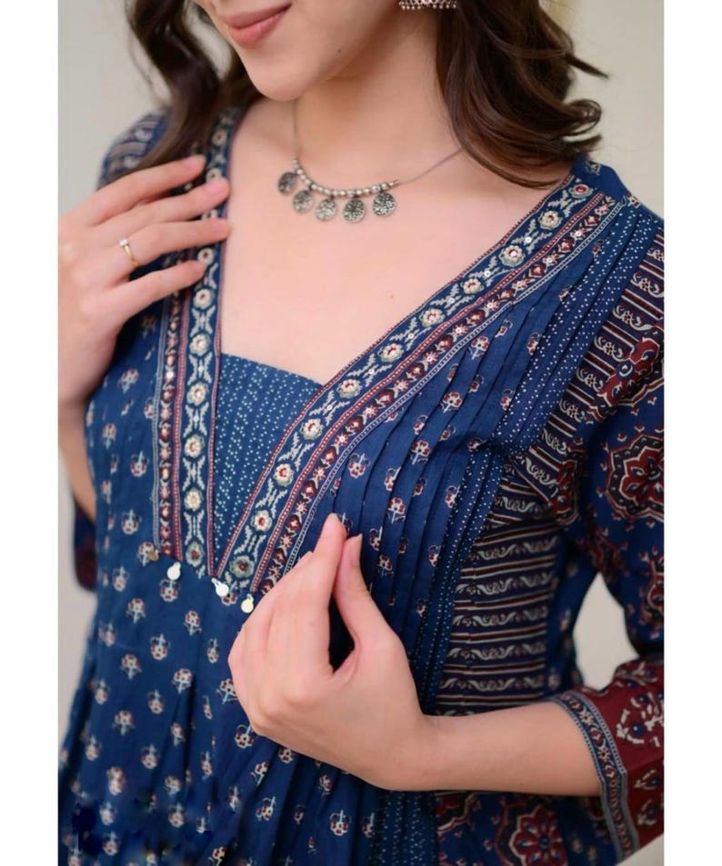 K&ouml;nnte beinhalten: Ein blau gemustertes Kleid mit V-Ausschnitt und Dreiviertel&auml;rmeln. Das Kleid hat ein detailliertes Design mit Falten und dekorativen Verzierungen. Eine silberne Halskette mit kreisf&ouml;rmigen Anh&auml;ngern wird mit dem Kleid getragen.