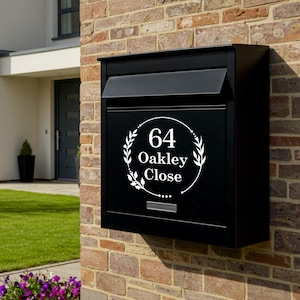 May include: A black mailbox mounted on a brick wall, with the address "64 Oakley Close" in a white circular design. The mailbox has a sloped top and a slot for mail. A modern house and green lawn are in the background.