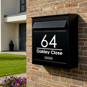 May include: A black mailbox mounted on a brick wall, displaying the number "64" and the text "Oakley Close" in white. The mailbox has a sloped top and a small slot for mail. The background shows a modern house with a green lawn and flowers.