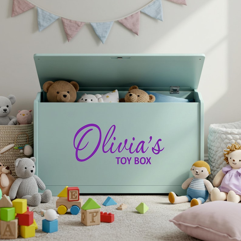 A purple personalised vinyl toy box name sticker reading 'Olivia's Toy Box' applied to a pastel green toy chest, surrounded by soft toys and blocks in a child's playroom, ideal custom toy box decal for smooth, flat surfaces.