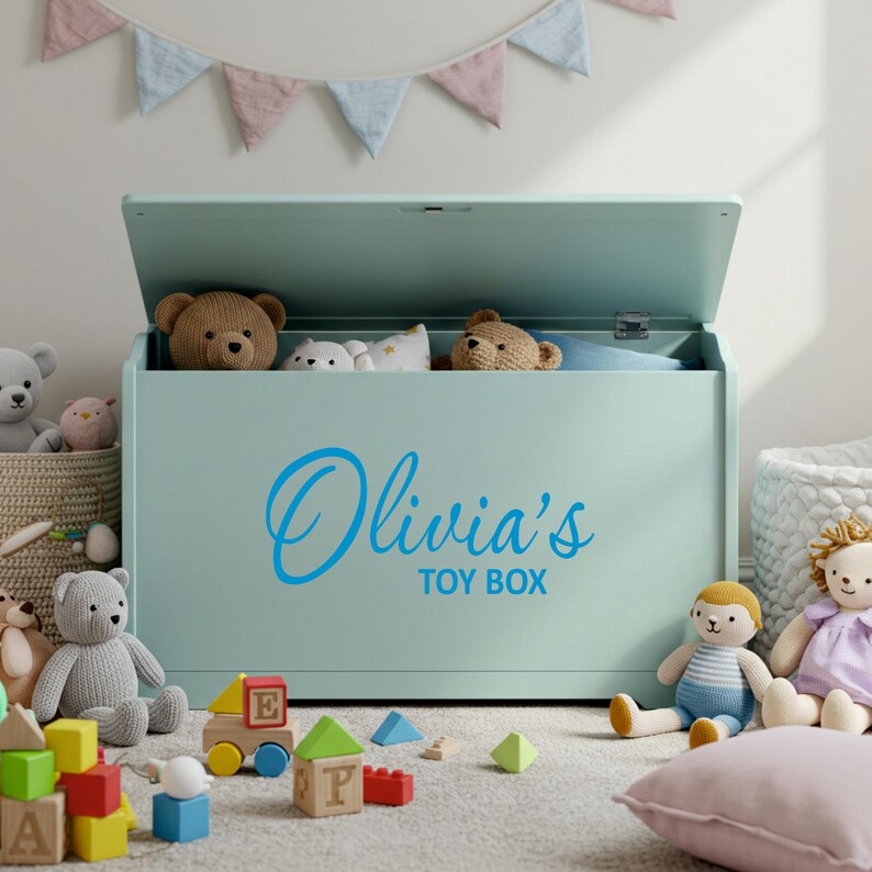 A blue personalised vinyl toy box name sticker reading 'Olivia's Toy Box' applied to a pastel green toy chest, surrounded by soft toys and blocks in a child's playroom, ideal custom toy box decal for smooth, flat surfaces.
