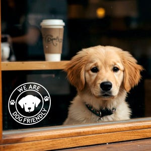 May include: A white sticker on a glass door with the text "We are dog friendly" in a circle with a paw print on each side and a silhouette of a dog's head in the centre. A golden retriever puppy is looking through the glass door.