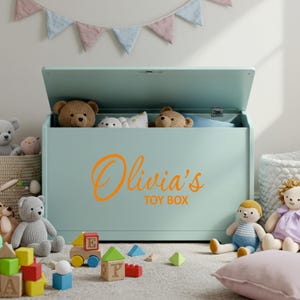 A orange personalised vinyl toy box name sticker reading 'Olivia's Toy Box' applied to a pastel green toy chest, surrounded by soft toys and blocks in a child's playroom, ideal custom toy box decal for smooth, flat surfaces.