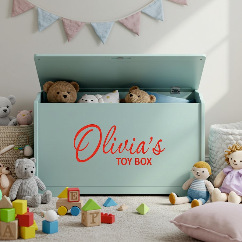 A red personalised vinyl toy box name sticker reading 'Olivia's Toy Box' applied to a pastel green toy chest, surrounded by soft toys and blocks in a child's playroom, ideal custom toy box decal for smooth, flat surfaces.