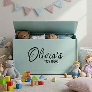 A black personalised vinyl toy box name sticker reading 'Olivia's Toy Box' applied to a pastel green toy chest, surrounded by soft toys and blocks in a child's playroom, ideal custom toy box decal for smooth, flat surfaces.