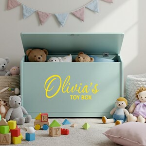 A yellow personalised vinyl toy box name sticker reading 'Olivia's Toy Box' applied to a pastel green toy chest, surrounded by soft toys and blocks in a child's playroom, ideal custom toy box decal for smooth, flat surfaces.