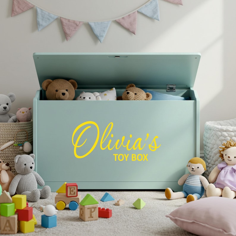A yellow personalised vinyl toy box name sticker reading 'Olivia's Toy Box' applied to a pastel green toy chest, surrounded by soft toys and blocks in a child's playroom, ideal custom toy box decal for smooth, flat surfaces.