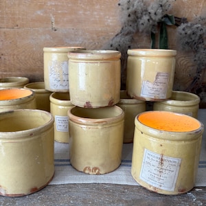 May include: A selection of antique, yellow ceramic jars with labels. The jars are cylindrical with a slightly flared rim. Some have labels with text. The interior of some jars is orange.