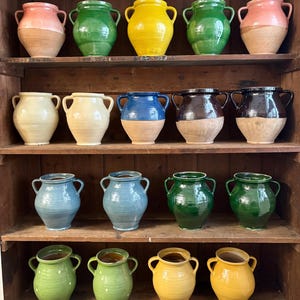 May include: A display of glazed ceramic vases in a variety of colours, including pink, green, yellow, and blue, arranged on wooden shelves. Each vase has two handles and a natural clay-coloured base. The vases are of varying sizes.