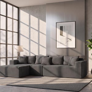 May include: A modern living room featuring a gray sectional sofa with several matching throw pillows. A framed abstract art piece hangs on the wall. A potted plant and a floor lamp are also present. Large windows allow sunlight to stream into the room.