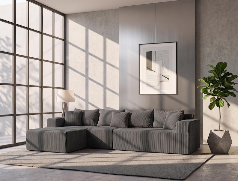 May include: A modern living room featuring a gray sectional sofa with several matching throw pillows. A framed abstract art piece hangs on the wall. A potted plant and a floor lamp are also present. Large windows allow sunlight to stream into the room.